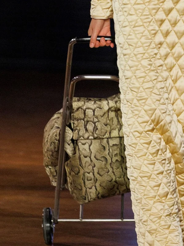 Closer: the hand on the trolley handle, a snake print bag on wheels, beside the diamond quilting of the dress.