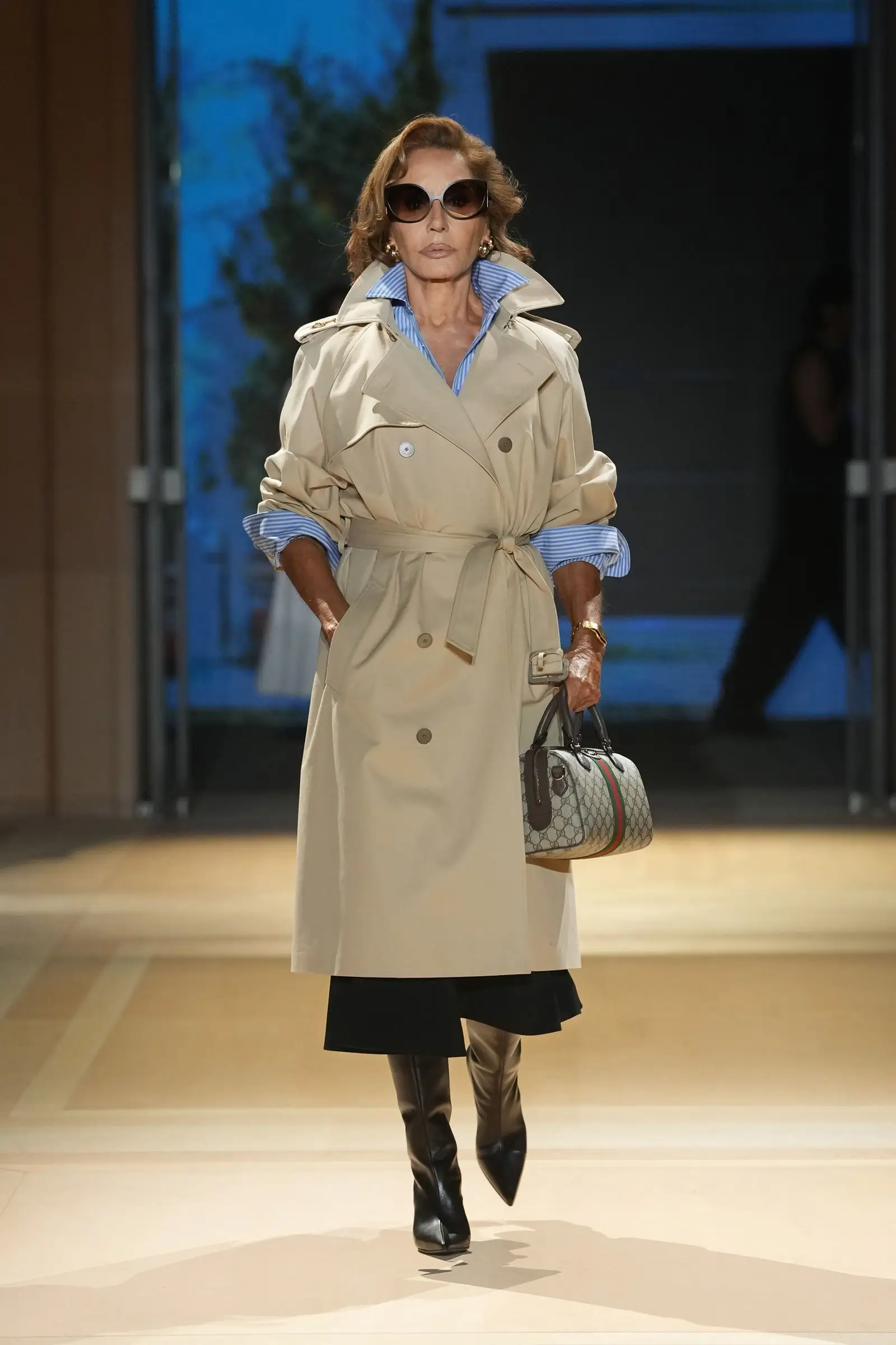 Gucci SS27, Look 10. Naty Abascal in a belted beige trench and dark sunglasses.
